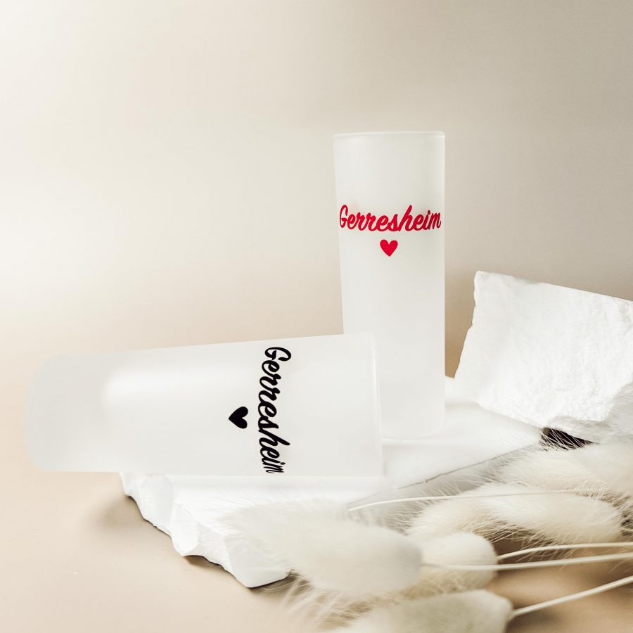 Zwei matte Shotgläser mit dem Schriftzug „Gerresheim“ und Herzmotiv, einmal in Schwarz und einmal in Rot, auf hellem terrazzoähnlichem Untergrund.