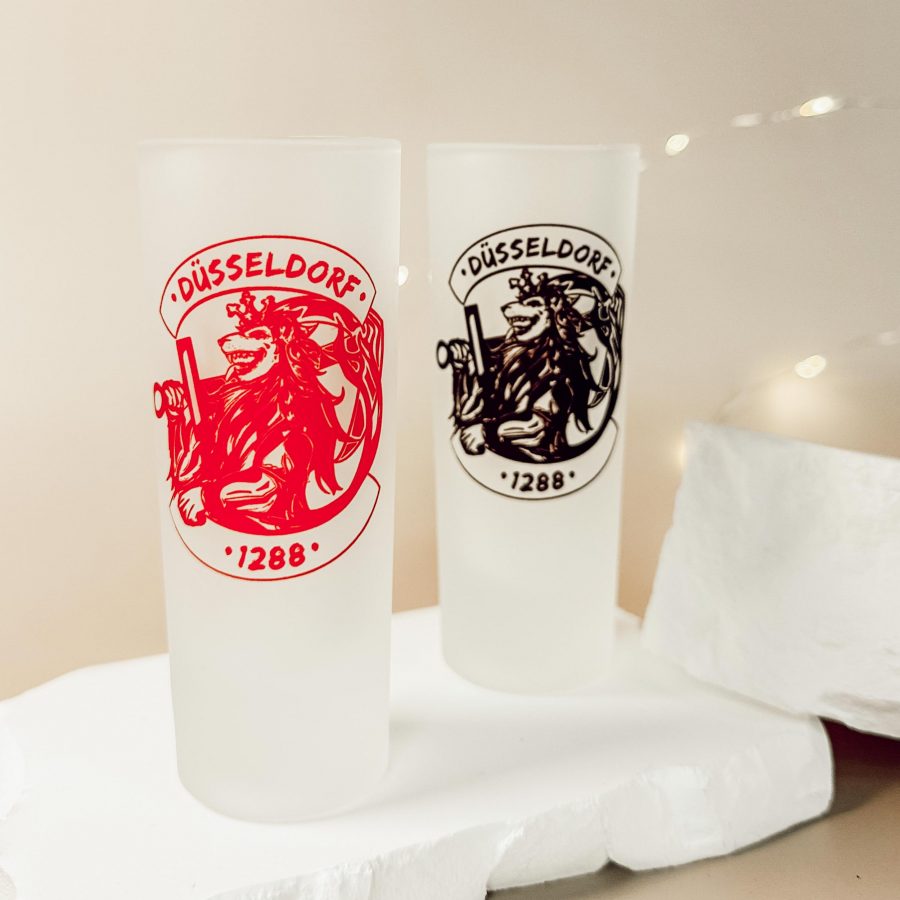 Zwei matte Shotgläser mit dem Düsseldorfer Löwenwappen „1288“, einmal in Rot und einmal in Schwarz, auf heller Deko-Fläche.