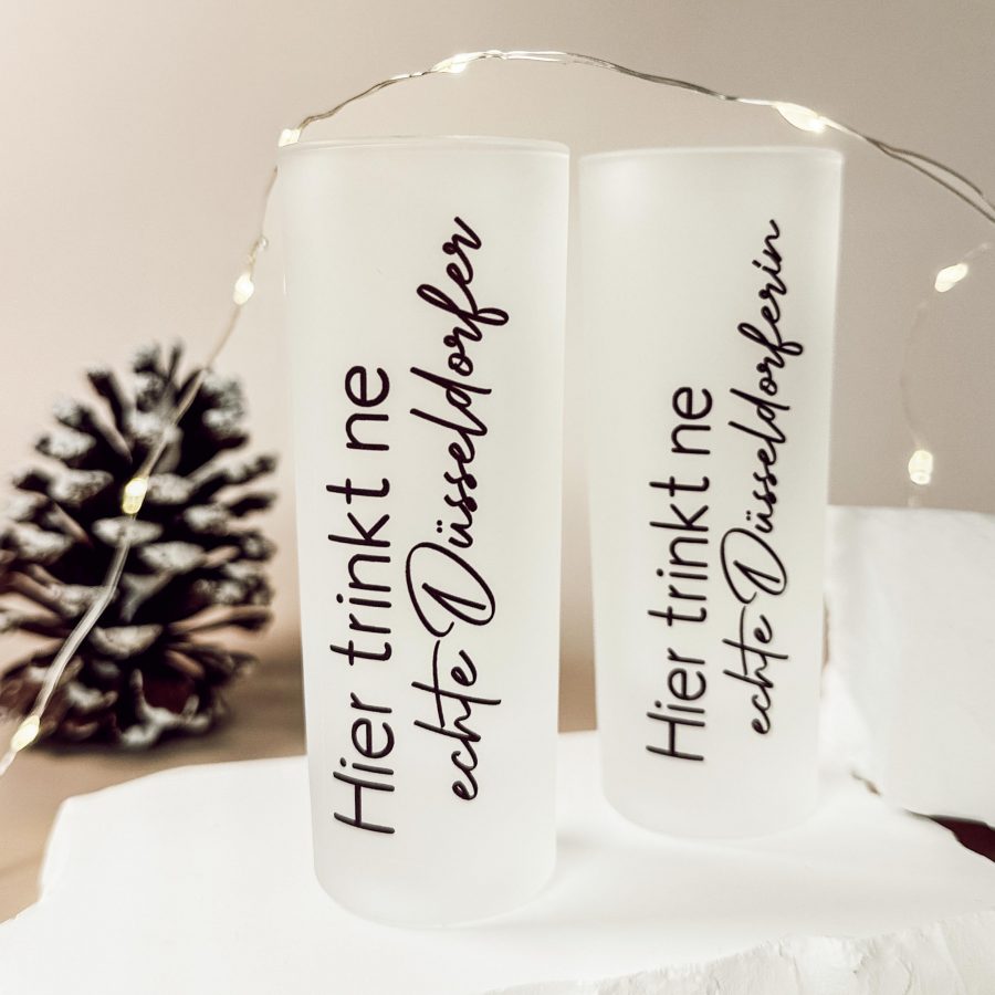 Zwei matte Shotgläser mit dem Aufdruck „Hier trinkt ne echte Düsseldorfer“ und „Hier trinkt ne echte Düsseldorferin“, dekorativ arrangiert mit Lichterkette und Tannenzapfen.